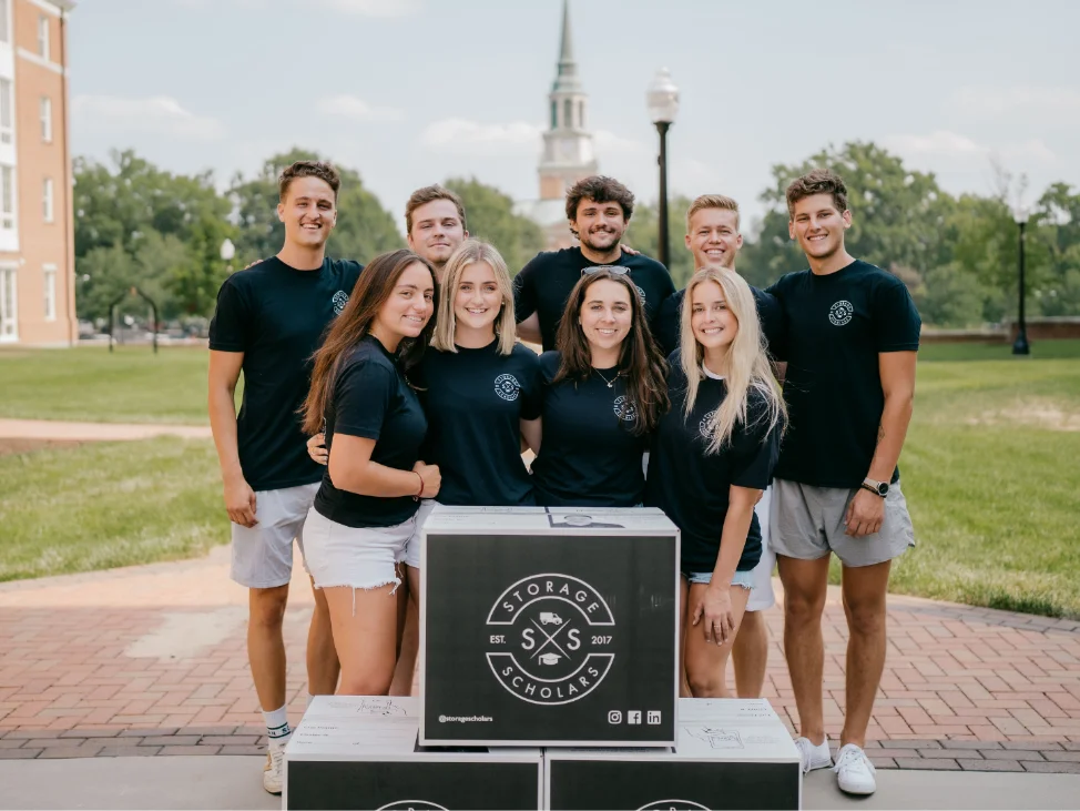 A crew of Storage Scholars students in matching shirts posing behind a stack of branded boxes on campus