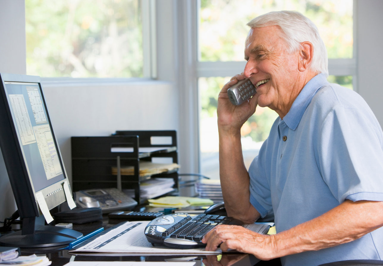 Older man sitting at a desk, speaking on landline phone while looking at a computer screen