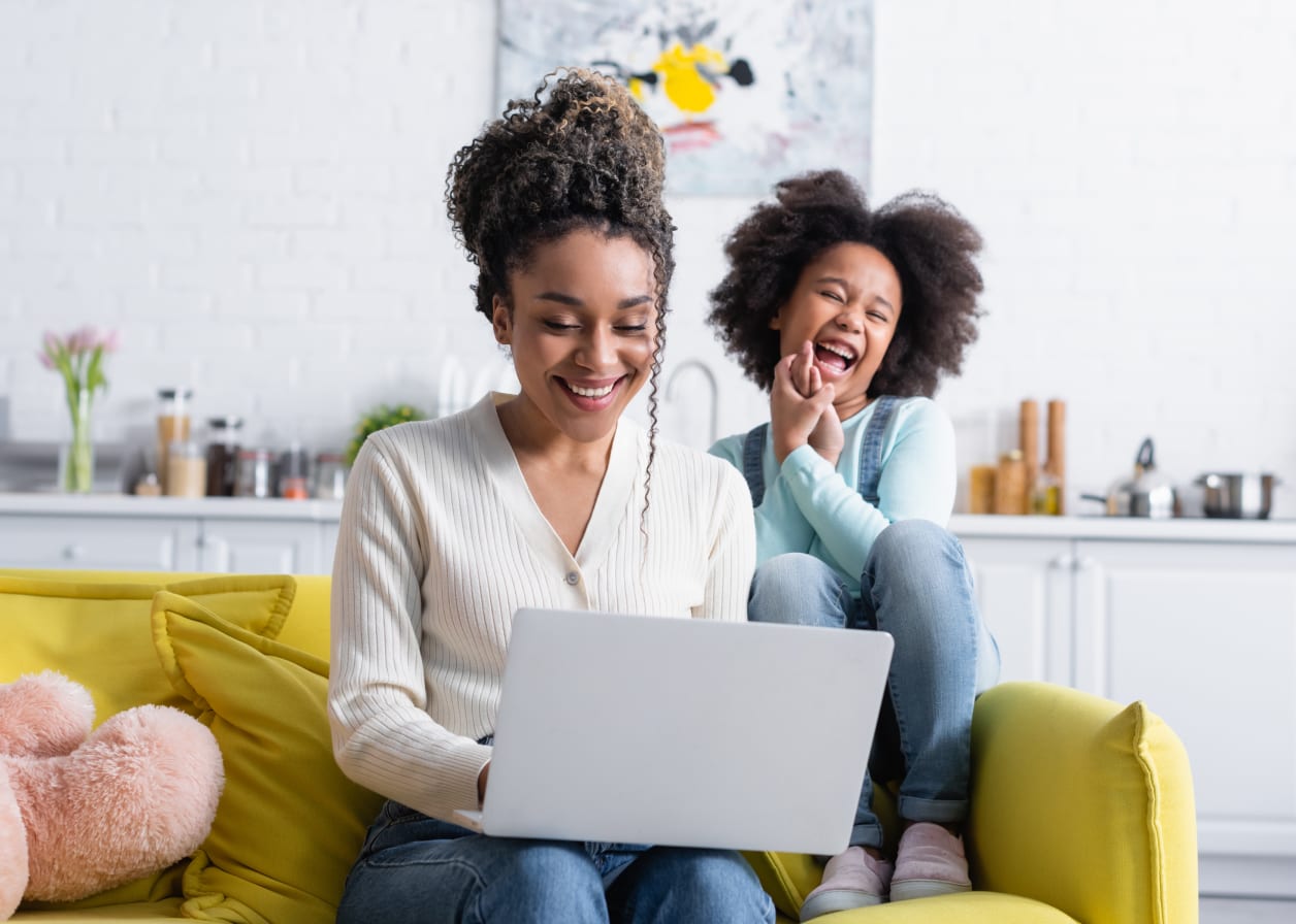 Mother and daughter sitting on the couch, laughing while using a laptop