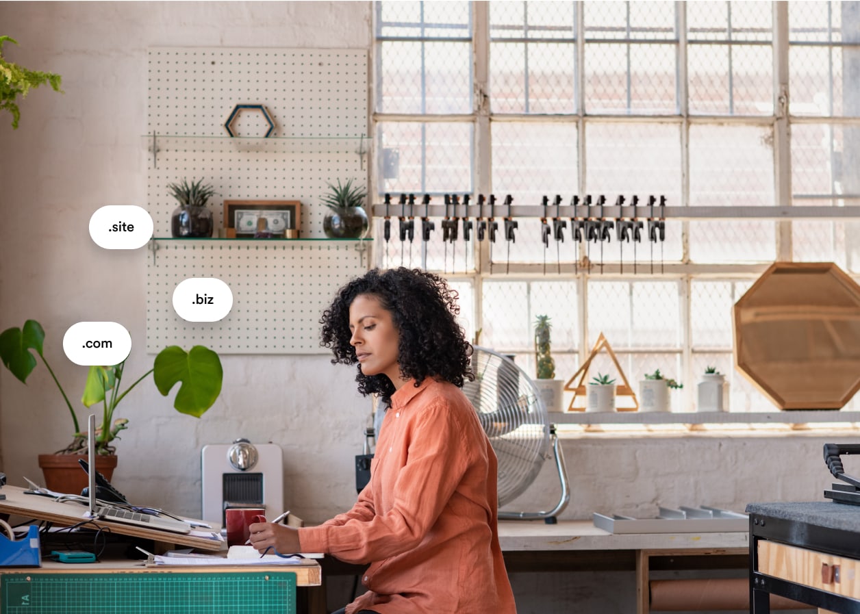 A young woman working on her laptop in her studio. Bubbles with '.com', '.site' and '.biz' float above her laptop. 