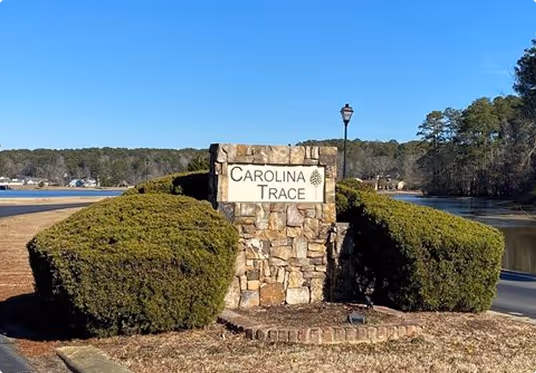 Stone sign reading "Carolina Trace" surrounded by trimmed bushes under a clear blue sky, with a lamp post nearby and a tranquil lake in the background.