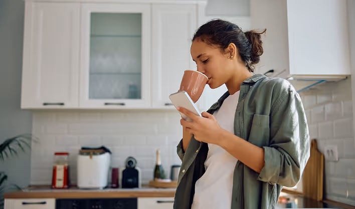Young woman in a casual outfit drinking from a mug while looking at her phone in a modern kitchen.