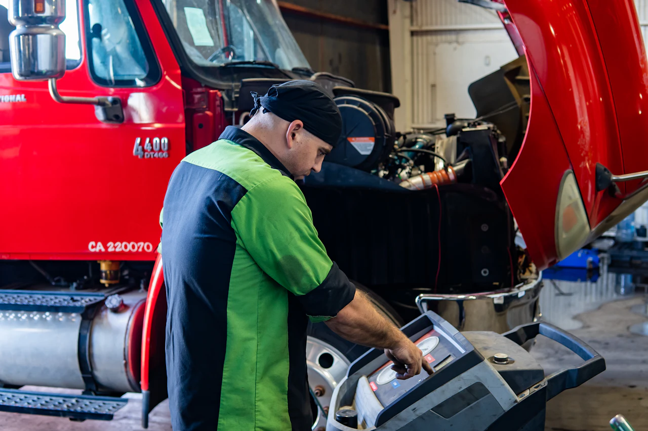 A mechanic wearing a black and green uniform operates diagnostic equipment in front of a red semi-truck with its hood lifted, exposing the engine. The scene takes place inside a well-lit garage, indicating a maintenance or repair procedure in progress.
