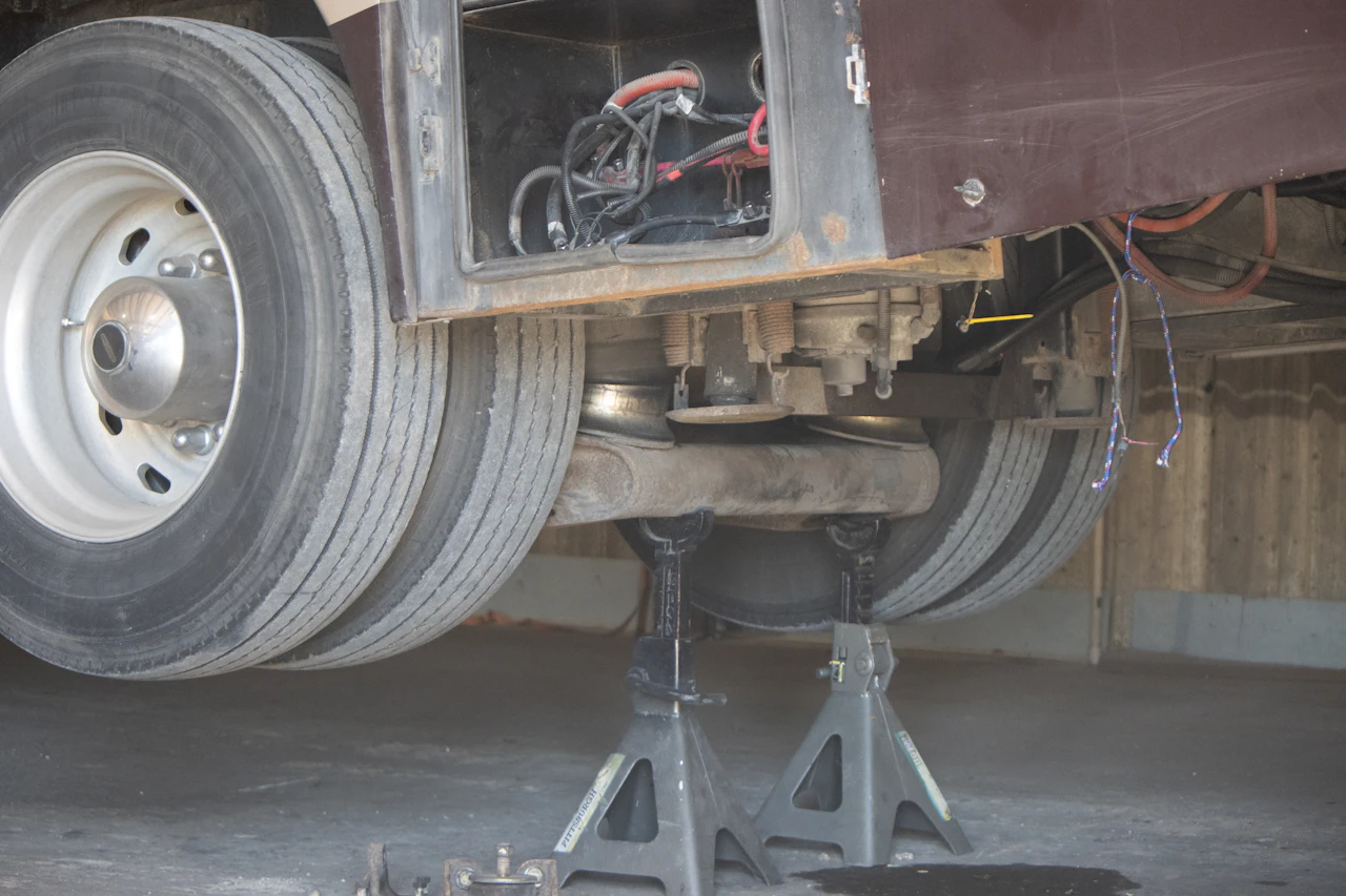 The rear axle of a large vehicle, likely a bus or RV, is lifted off the ground and supported by two heavy-duty jack stands, with its dual rear tires clearly visible. An open compartment reveals exposed electrical wiring above the axle.