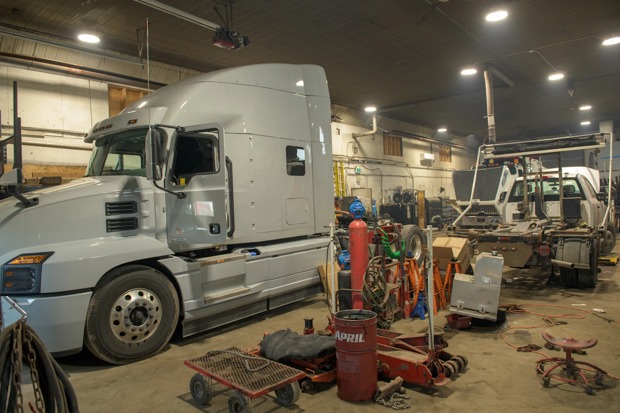A large silver semi-truck and a partially disassembled utility truck are parked inside a dimly lit workshop filled with industrial tools, gas cylinders, and scattered maintenance equipment. The scene suggests ongoing repair work in a busy garage setting.