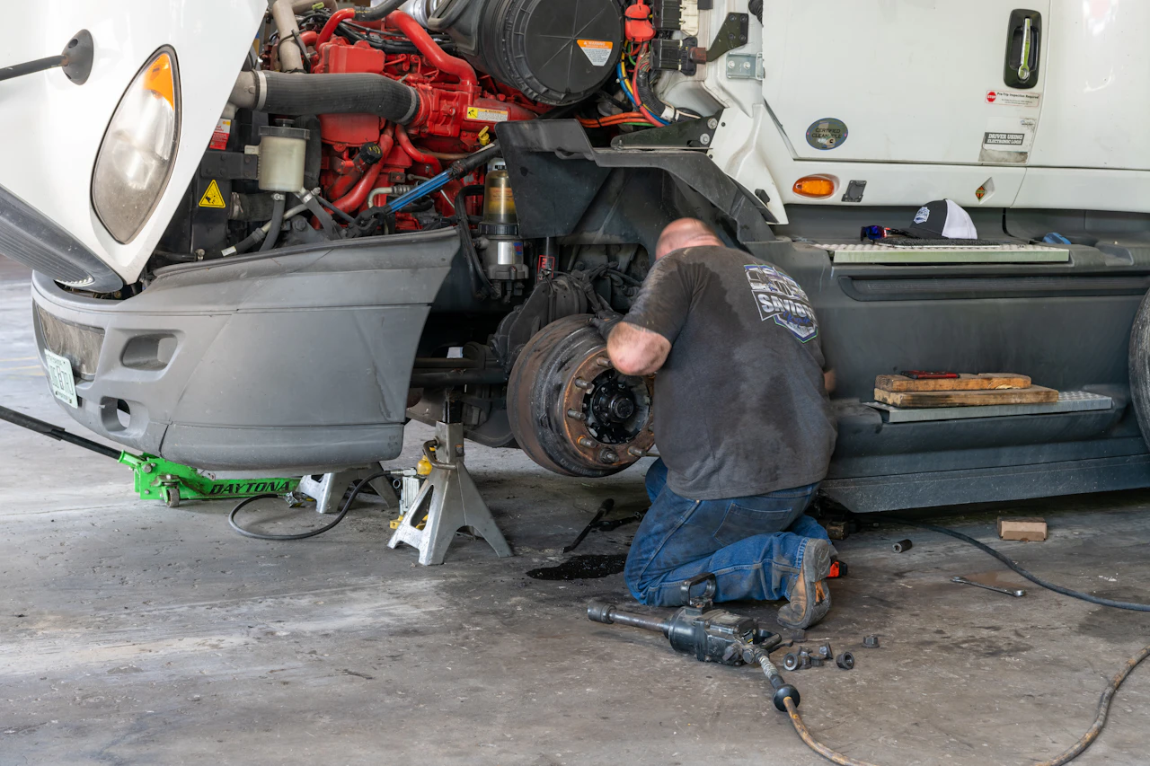 A mechanic kneels on the ground repairing the front wheel assembly of a white semi-truck propped up on jack stands. Tools, bolts, and an air impact wrench are scattered on the concrete floor nearby.