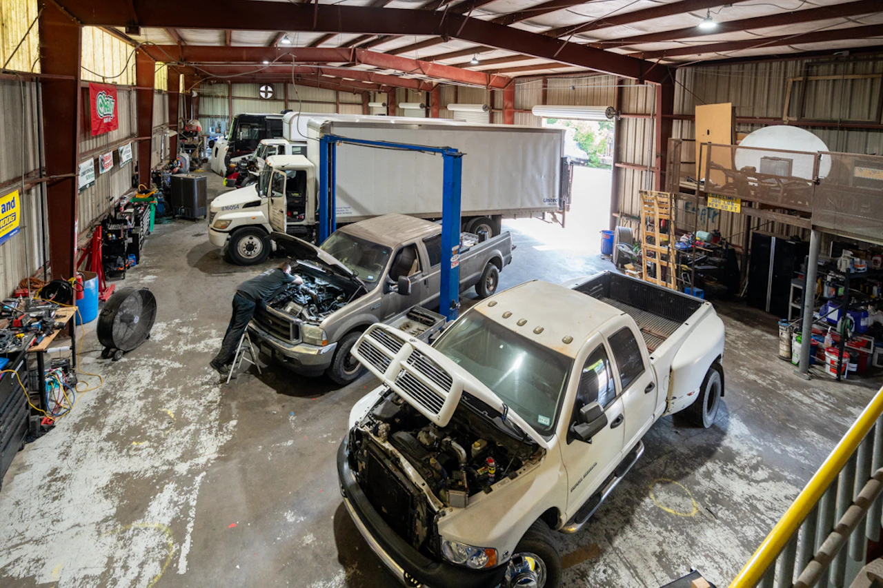 Diesel repair shop with multiple trucks in service bays, hoods open, and a technician working under bright overhead lighting.