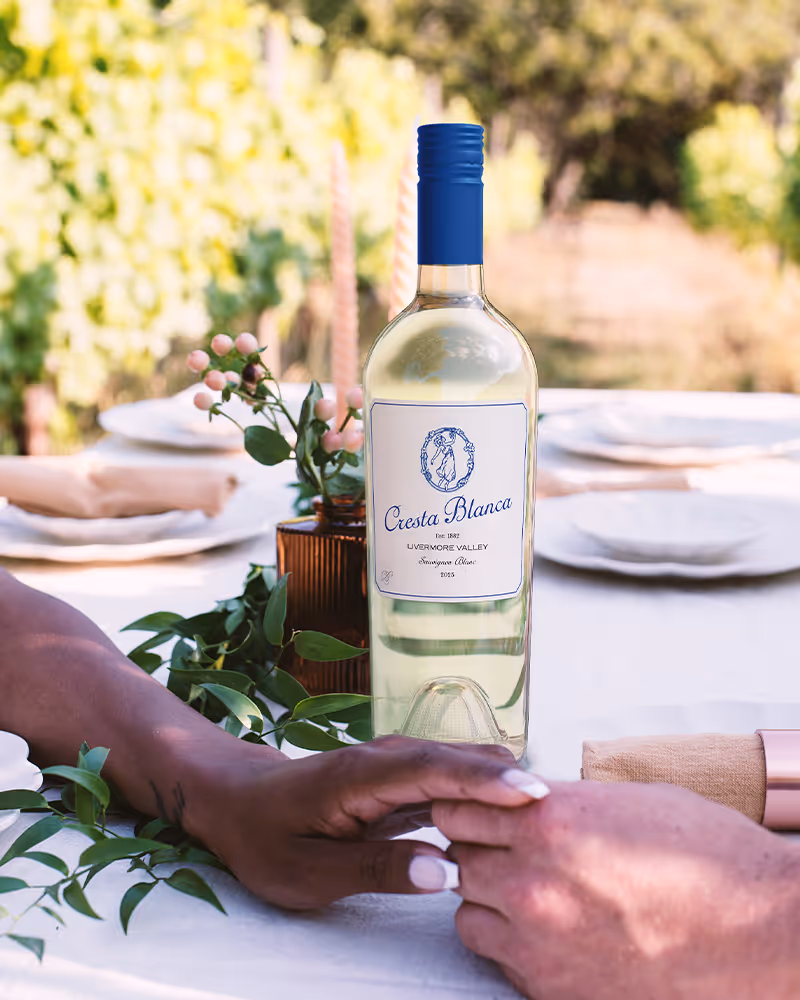 Two people holding hands on a table with a bottle of Cresta Blanca white wine and floral decor.