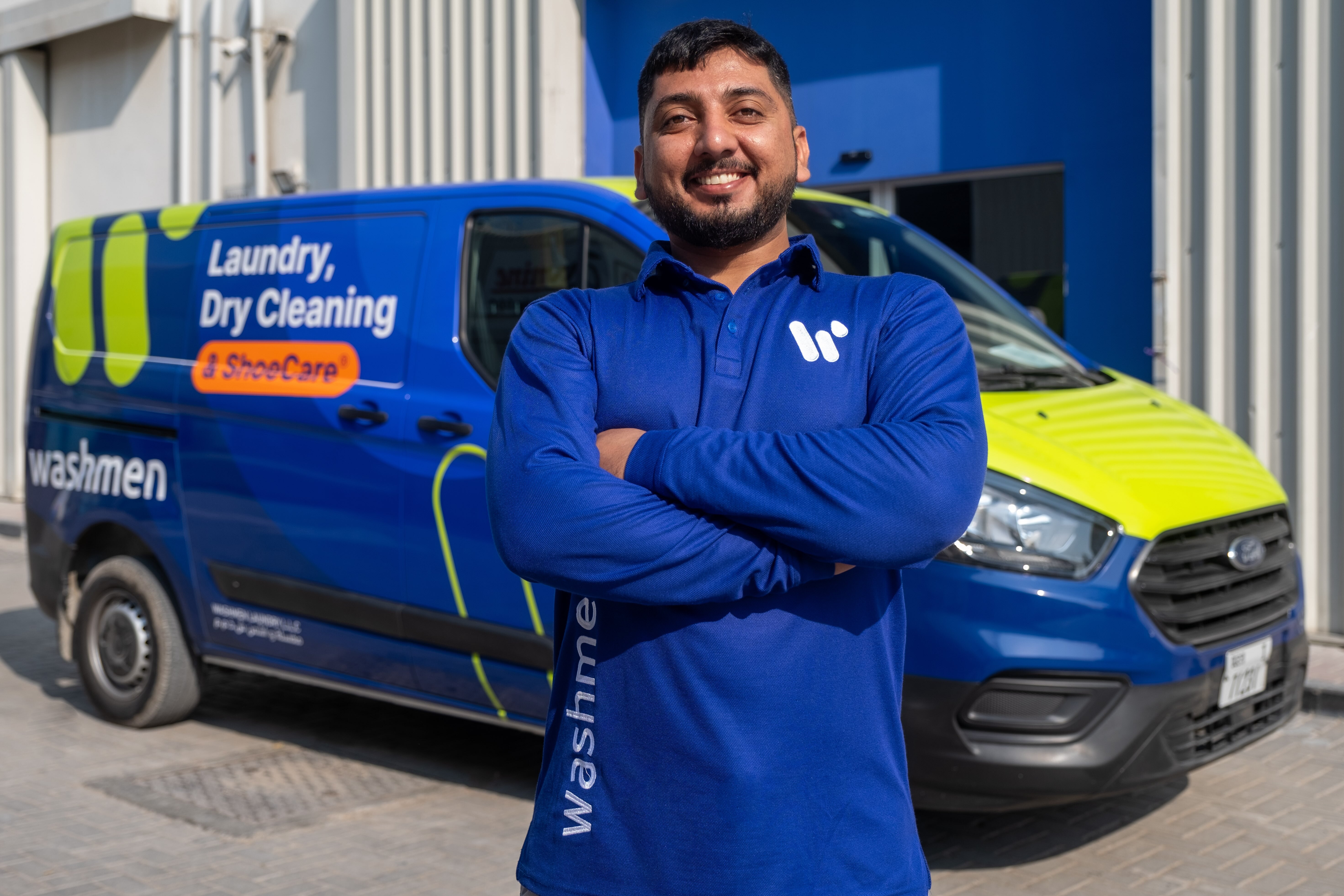 Smiling man in blue Washmen uniform standing with arms crossed in front of a blue and green Washmen service van.