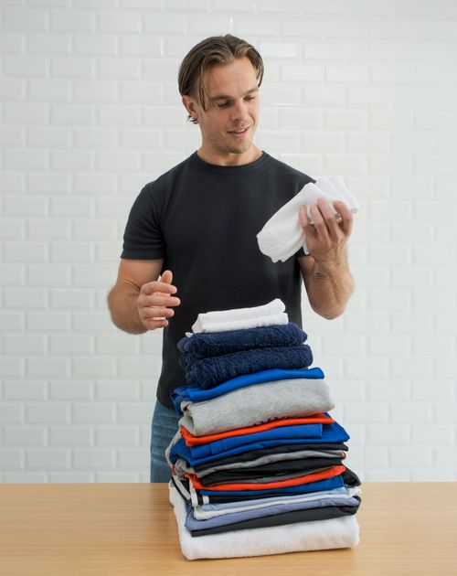 Man in black t-shirt folding a stack of neatly folded clothes on a wooden table.