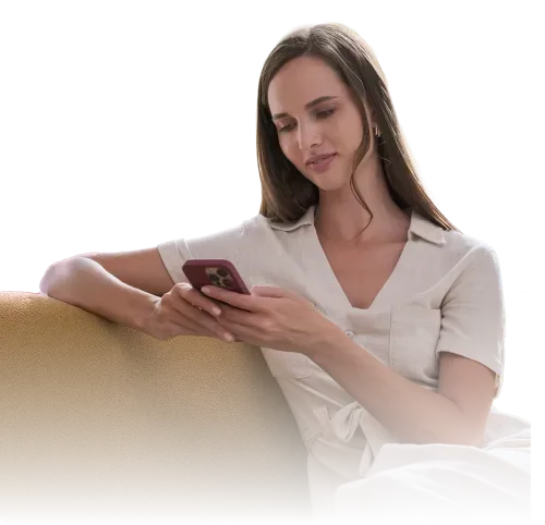 A woman sitting on a yellow couch, looking at her smartphone with a slight smile. She is wearing a light-colored, short-sleeved dress and has long hair styled down.