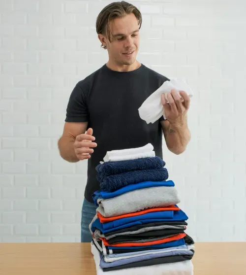Man in black shirt folding white socks next to a tall stack of folded clothes on a wooden table.