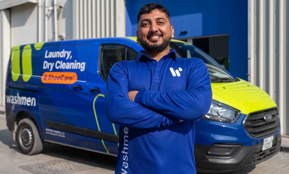 Мужчина в синей рубашке-поло держит костюм, закрытый пластиковым пакетом для одежды. Он стоит на фиолетовом фоне, демонстрируя дружелюбное поведение, и держит в руках белую сумку с надписью "стиральная машина".