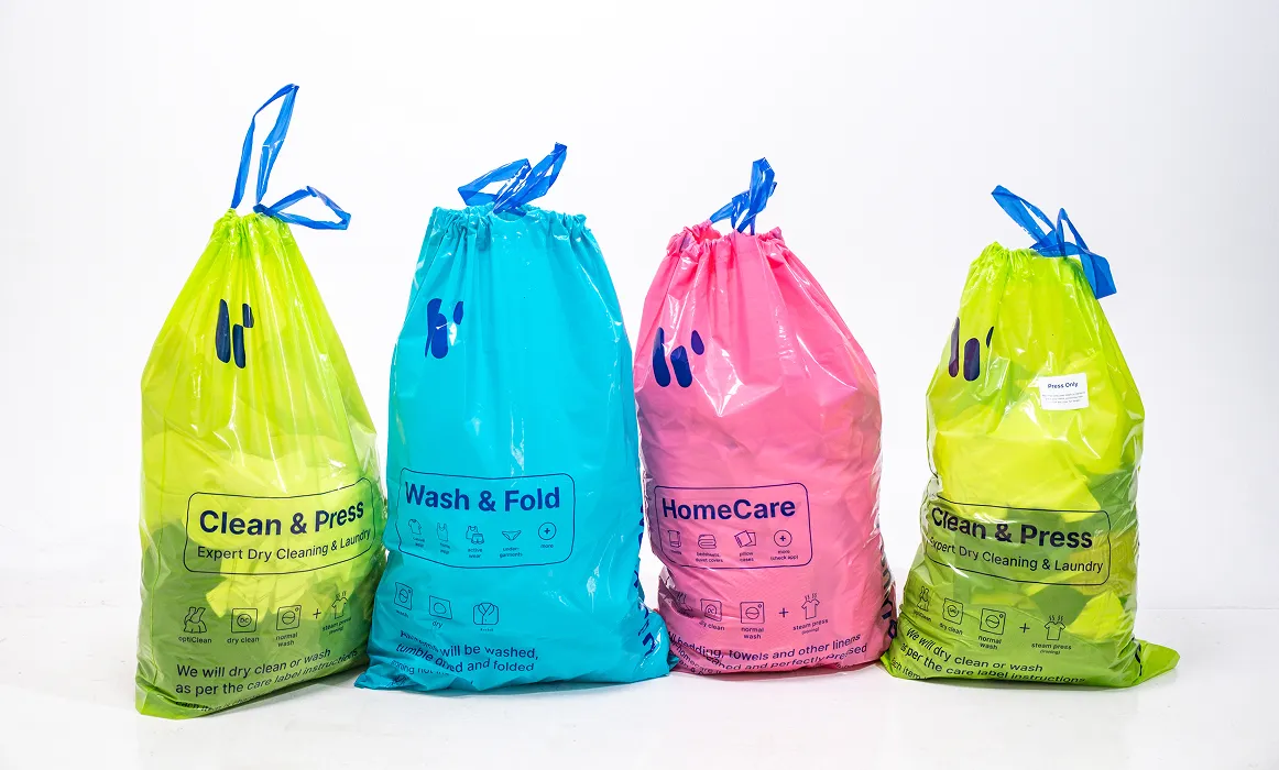 Женщина сидит на желтом диване и с легкой улыбкой смотрит на свой смартфон. На ней светлое платье с короткими рукавами, а длинные волосы уложены в прическу.