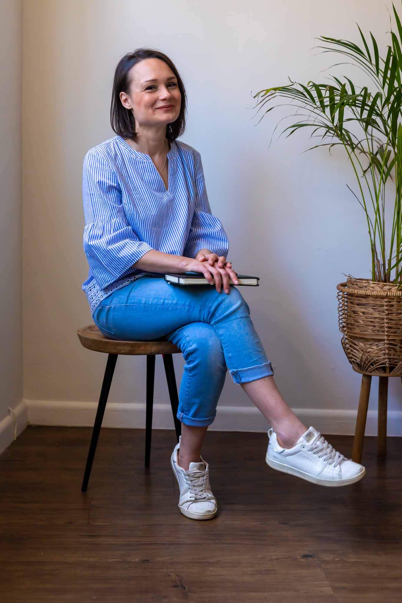 A woman with shoulder length black hair wearing a denim shirt and jeans sits cross-legged on a stool in the corner of a white room. She's smiling at the camera while holding a black notebook and pen.