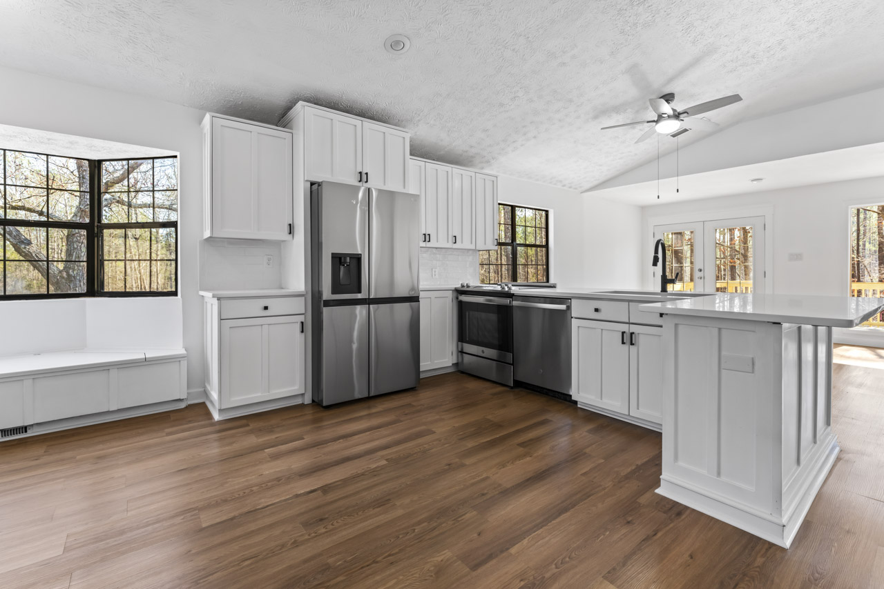 Bright modern kitchen with white cabinets, stainless steel appliances, wooden floor, and large windows showing trees outside.