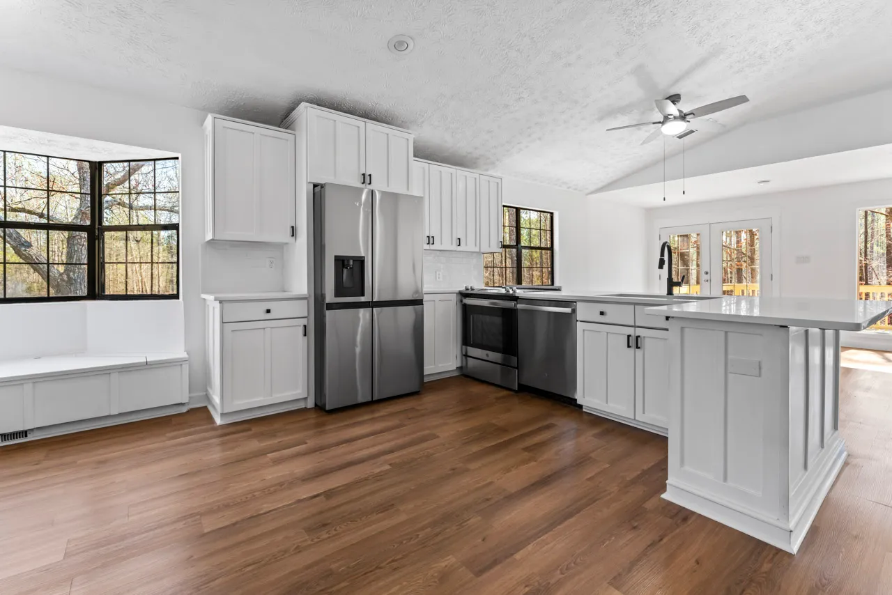 Bright modern kitchen with white cabinets, stainless steel appliances, wooden floor, and large windows showing trees outside.