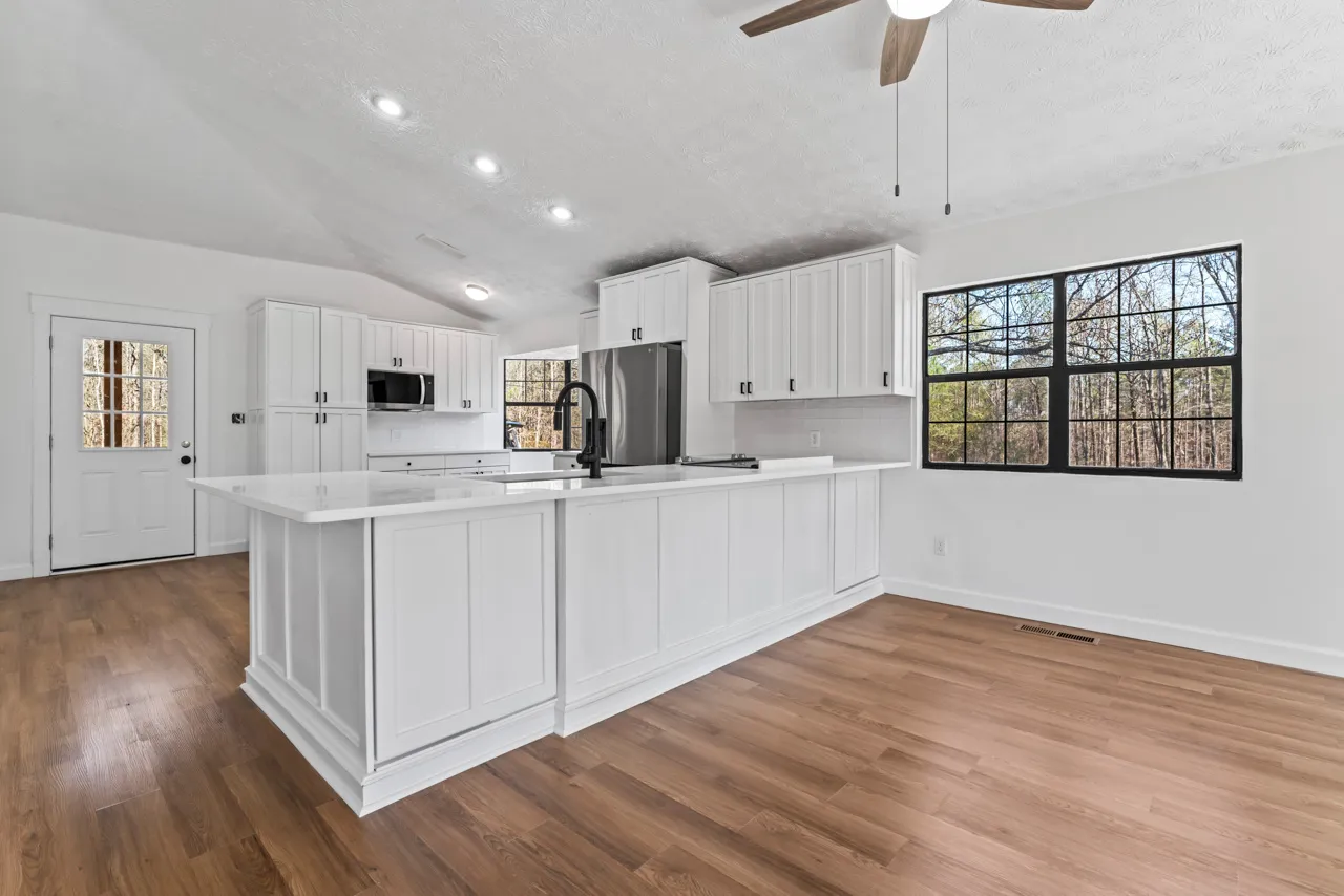 Bright kitchen with white cabinets, a large island with a black faucet, stainless steel refrigerator, wooden floor, and large windows showing trees outside.
