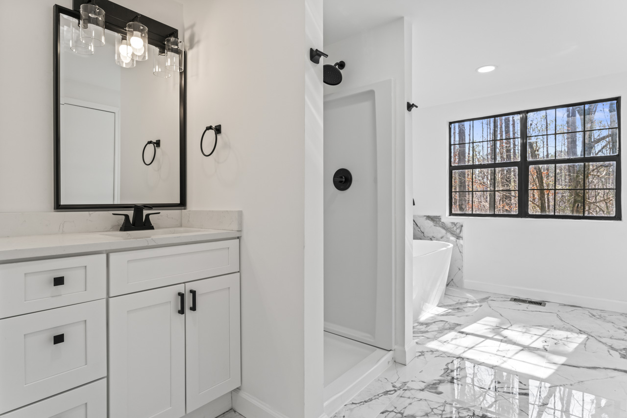 Bright bathroom with white vanity, black fixtures, walk-in shower, and large window showing trees outside.