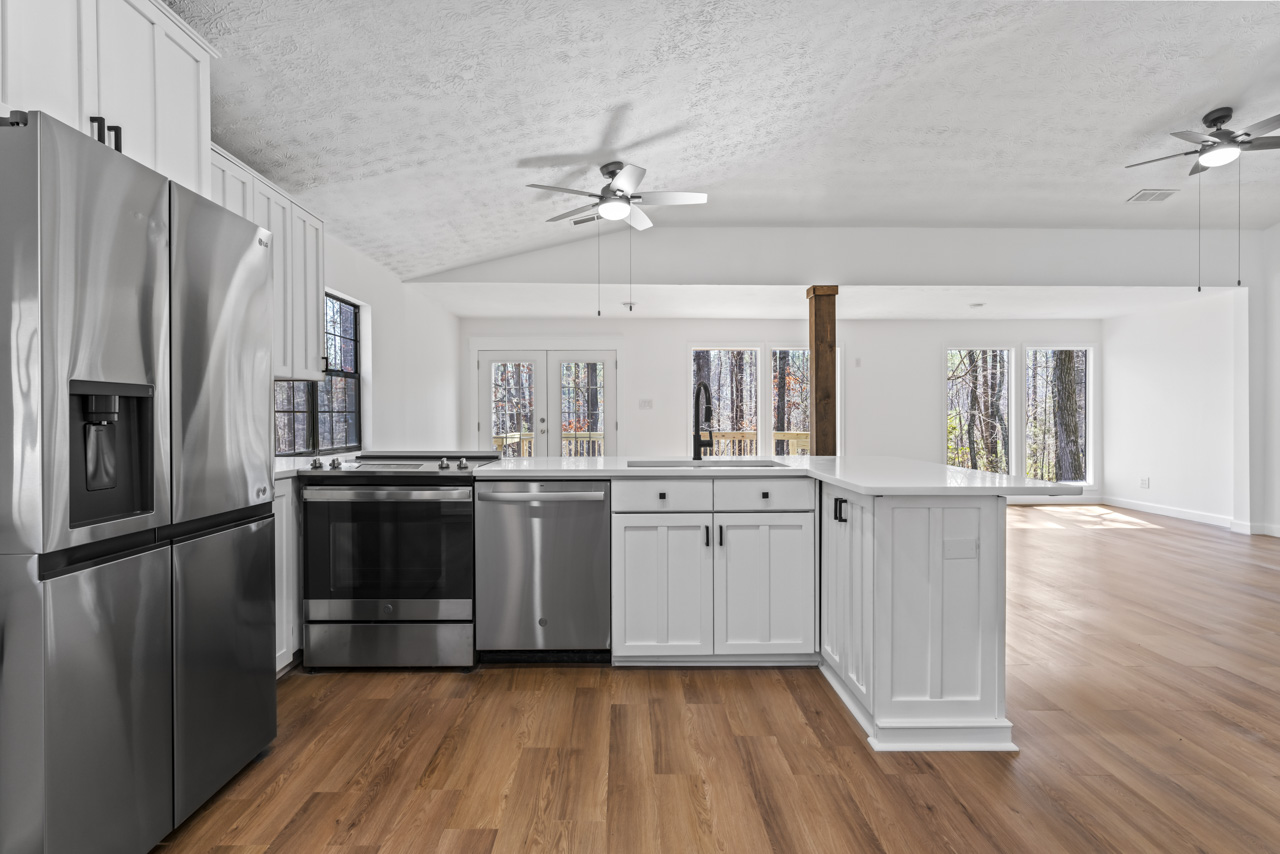 Modern kitchen with stainless steel refrigerator, stove, dishwasher, white cabinets, wood flooring, and ceiling fans, opening to a bright living area with large windows.