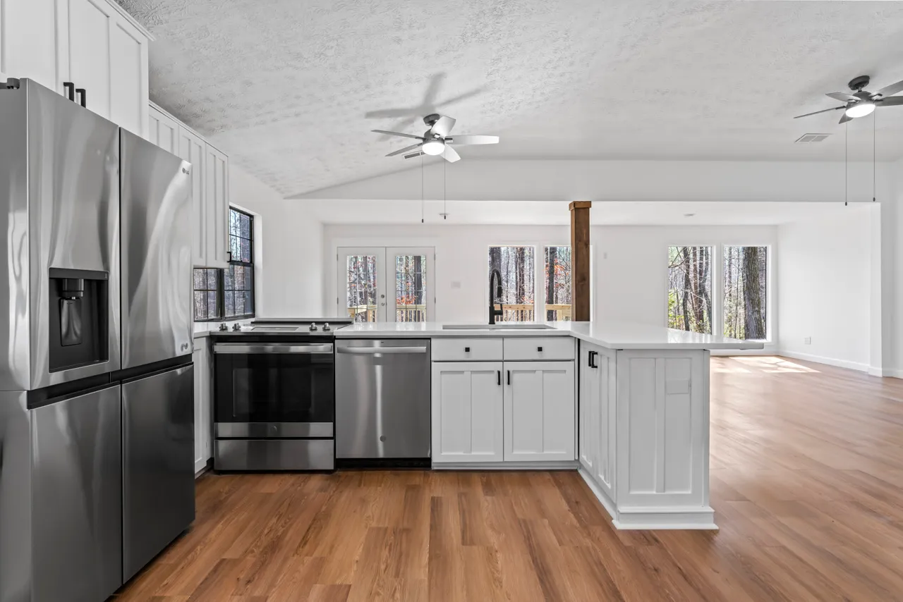Modern kitchen with stainless steel refrigerator, stove, dishwasher, white cabinets, wood flooring, and ceiling fans, opening to a bright living area with large windows.