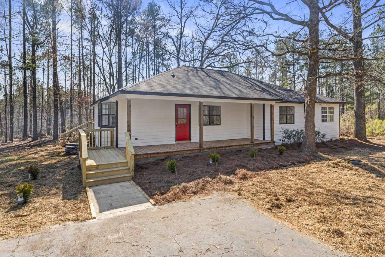Single-story white house with a gray roof, red front door, wooden porch, and surrounding bare trees under a clear blue sky.