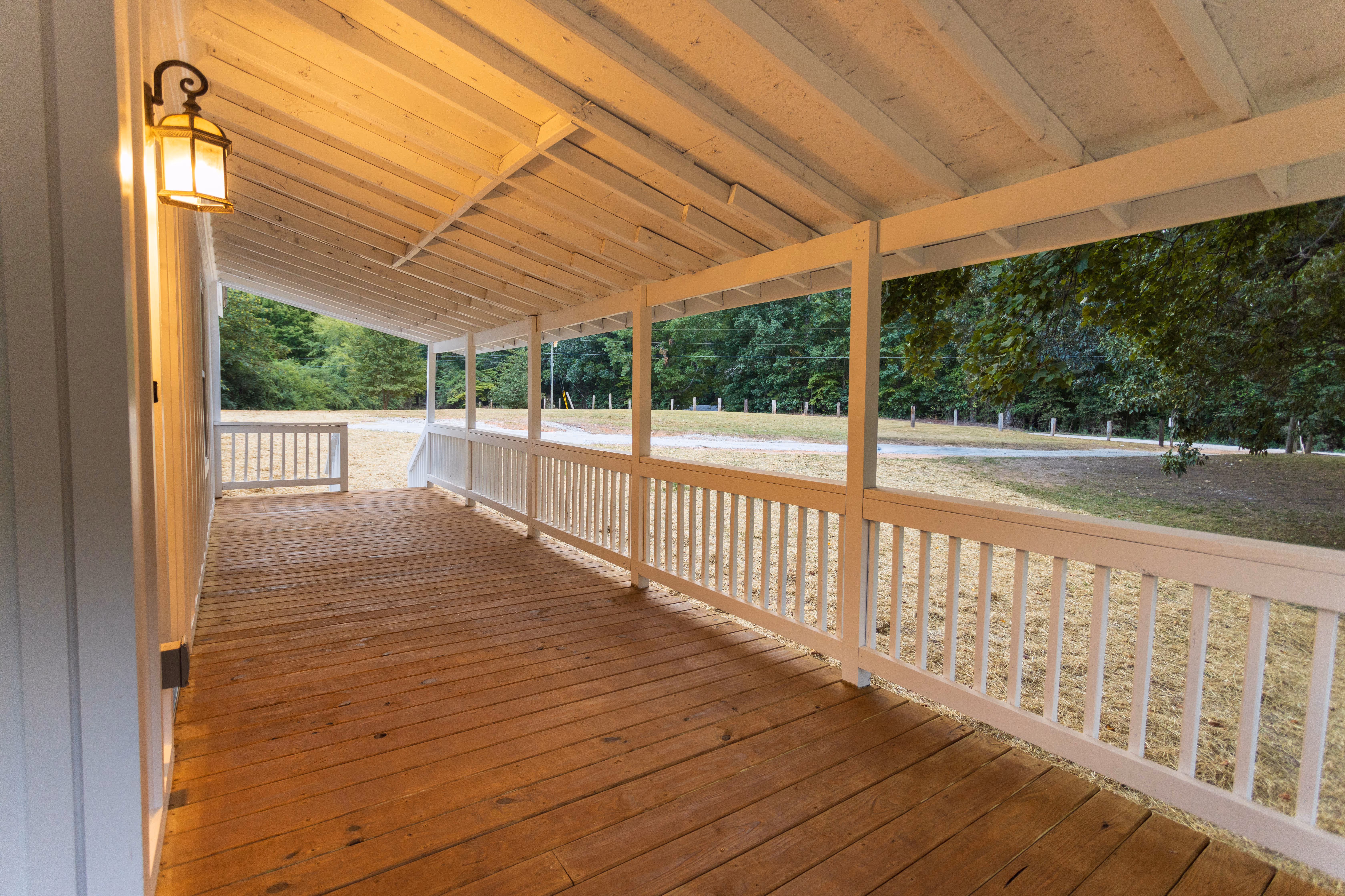 Wide covered wooden porch with white railings overlooking a grassy area and trees.