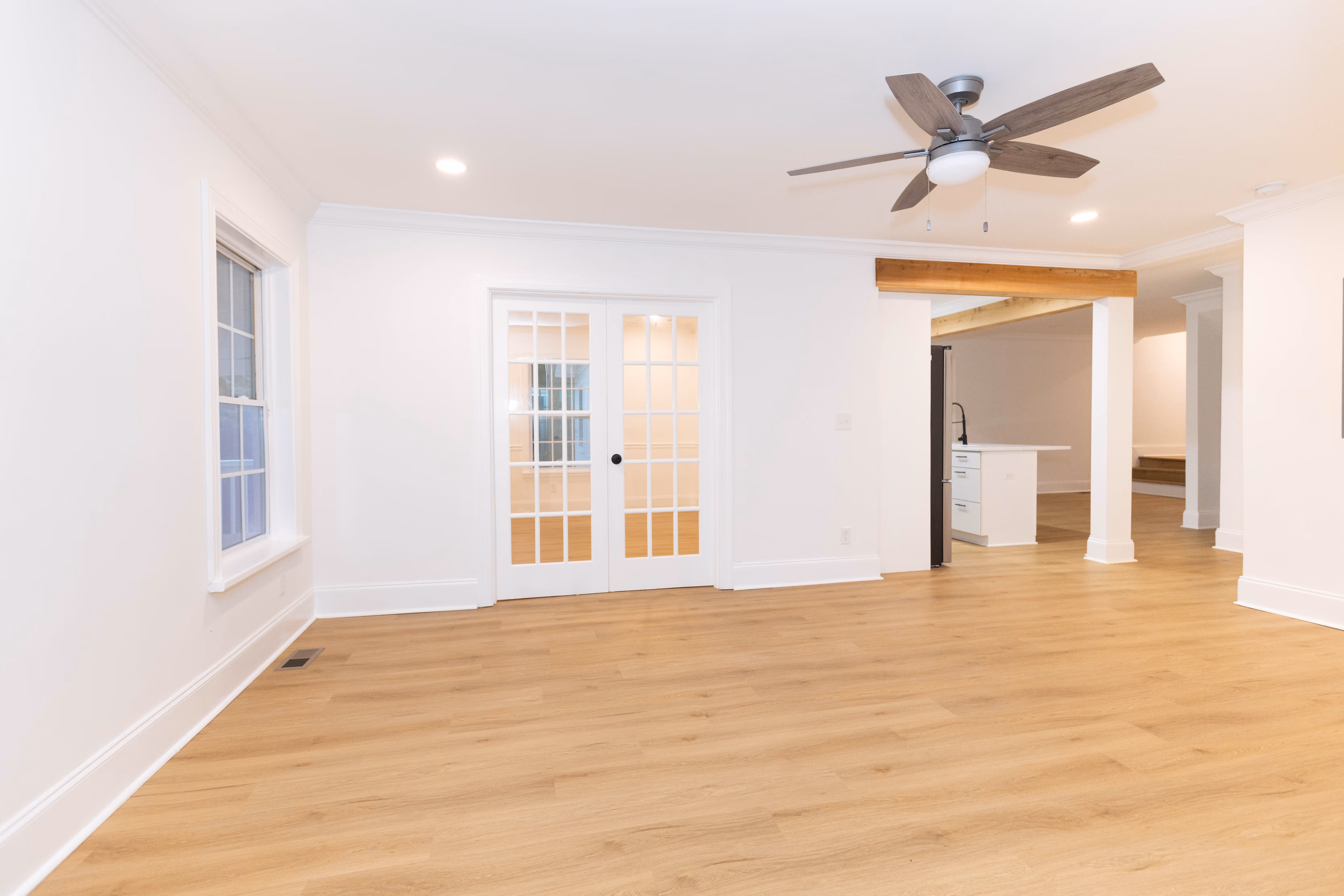 Bright empty room with light wood flooring, white walls, glass-paneled double doors, ceiling fan, and an open doorway to a kitchen area.