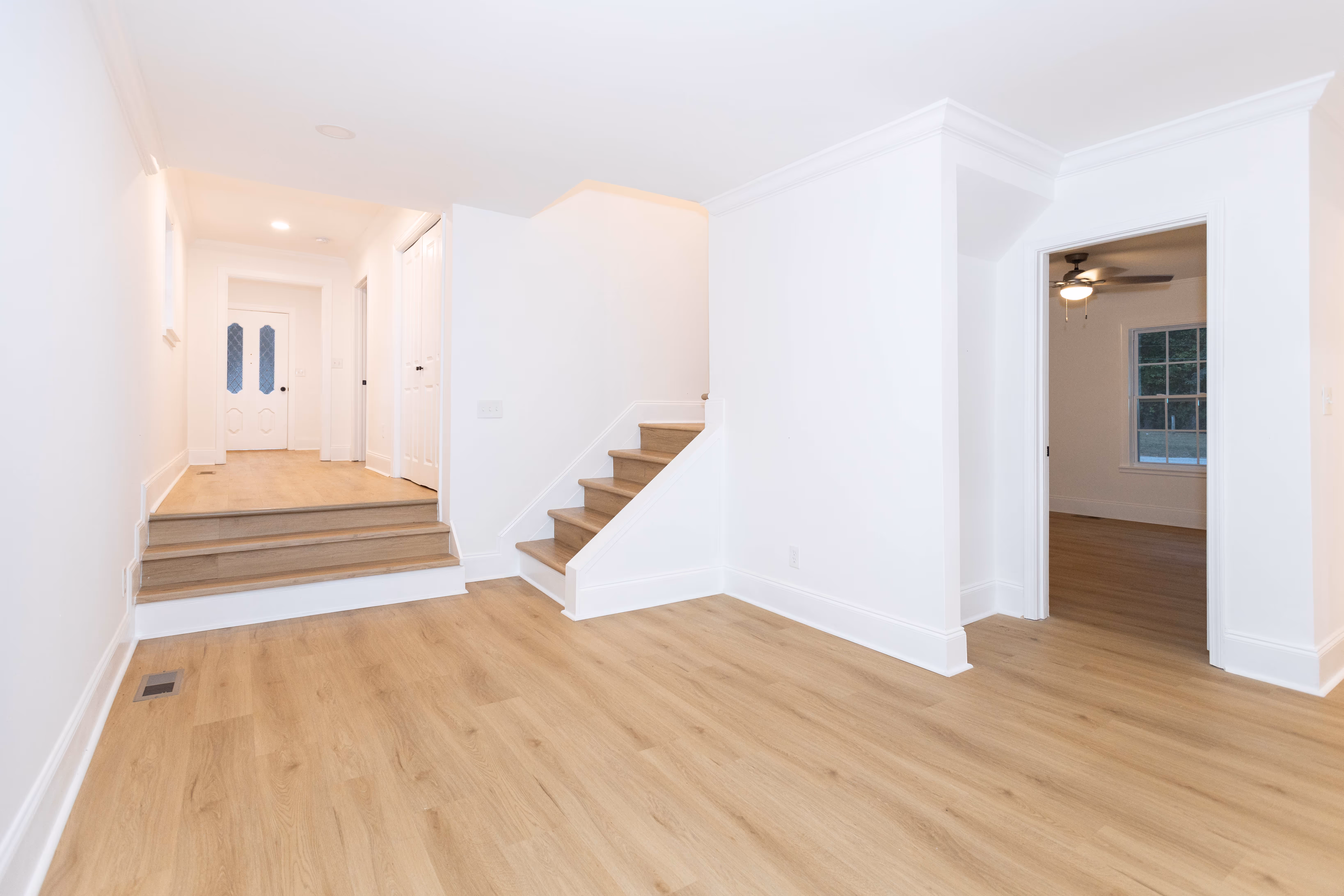 Bright interior with light wood flooring, white walls, stairs, hallway with white doors, and a room with a ceiling fan and window.
