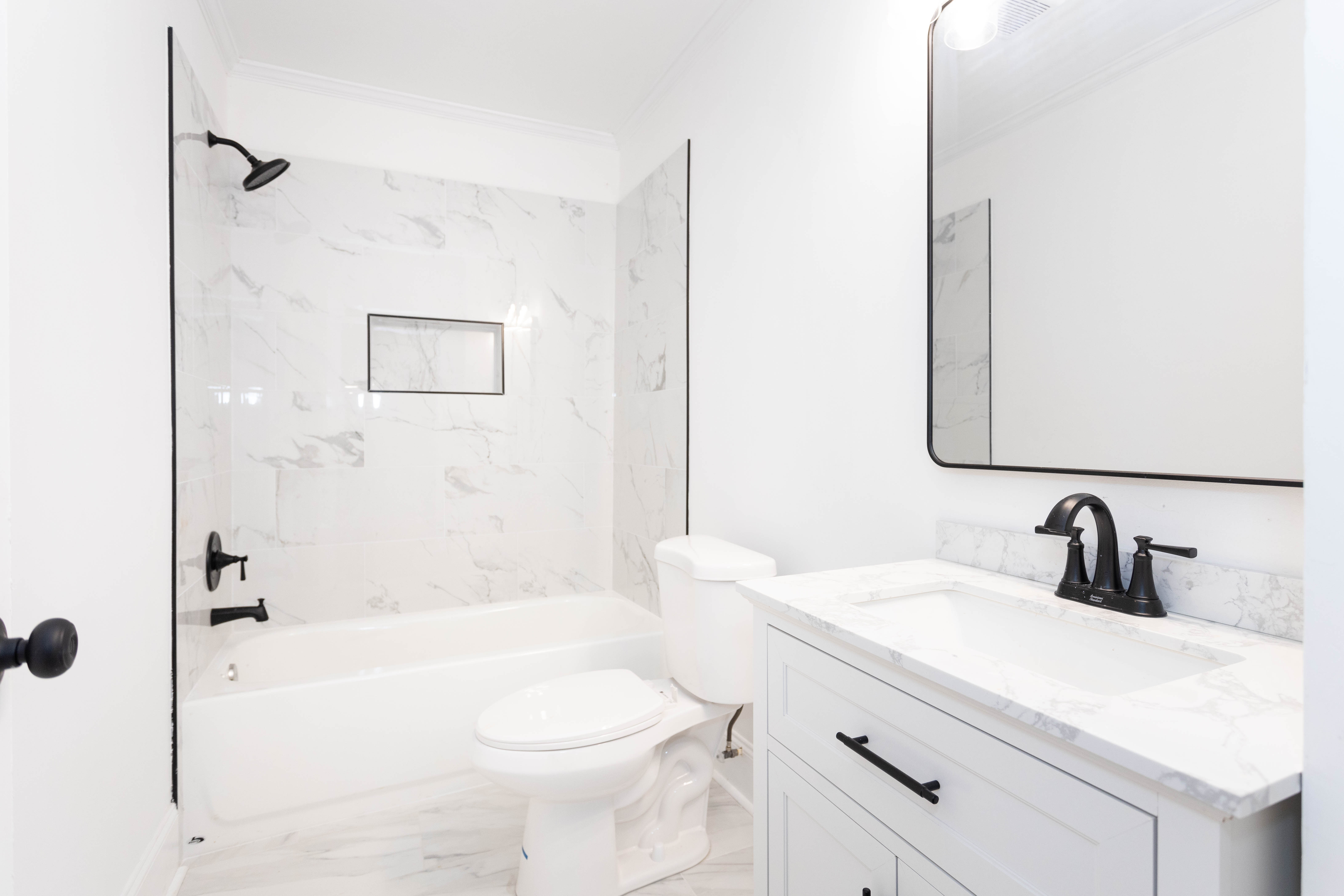 Bright white bathroom with marble tiled shower and bathtub, white toilet, and white vanity with black faucet and mirror.