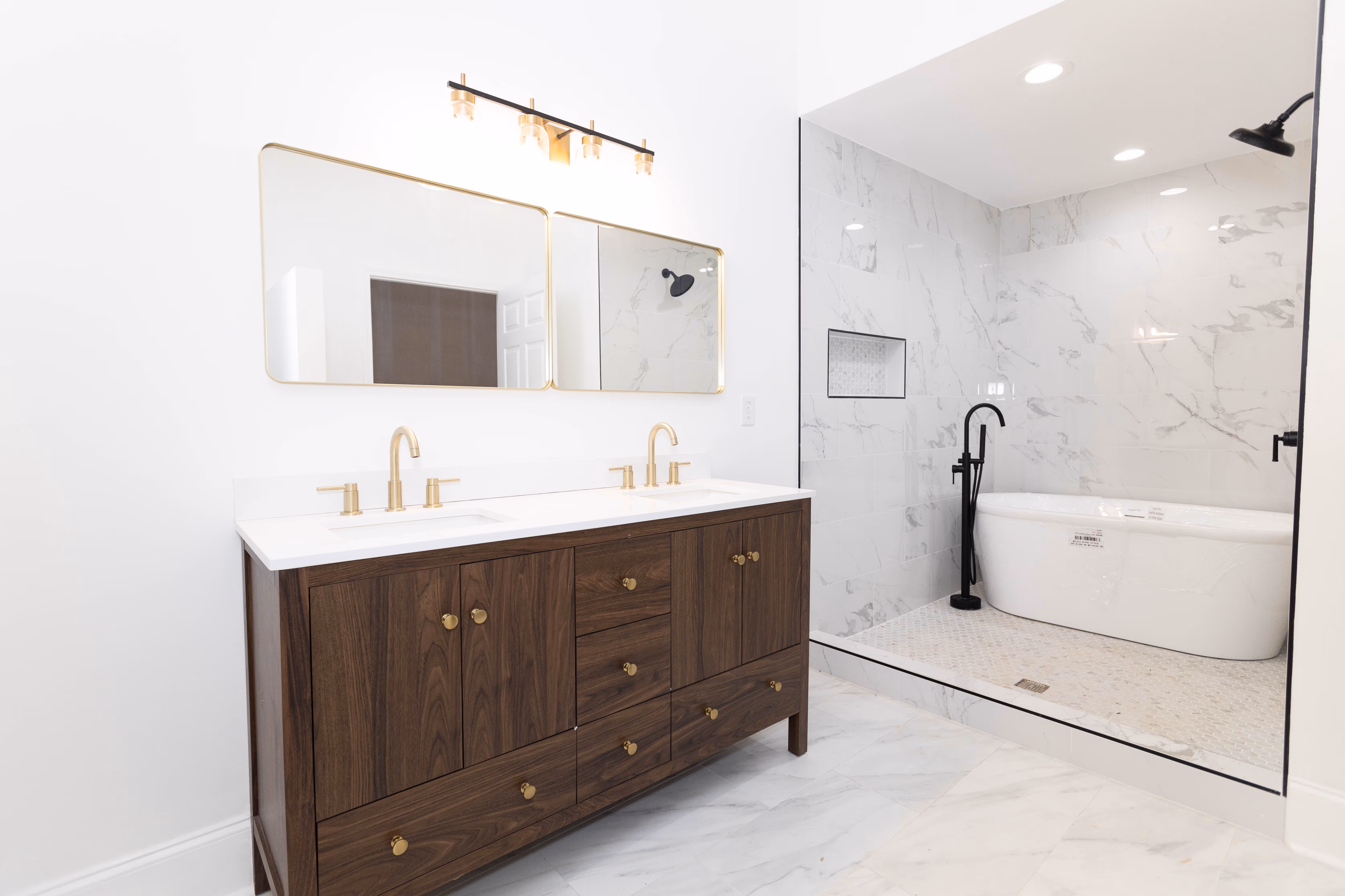 Modern bathroom with a dark wood double vanity with gold faucets and two mirrors, next to a glass-enclosed shower area featuring a freestanding bathtub and black fixtures.