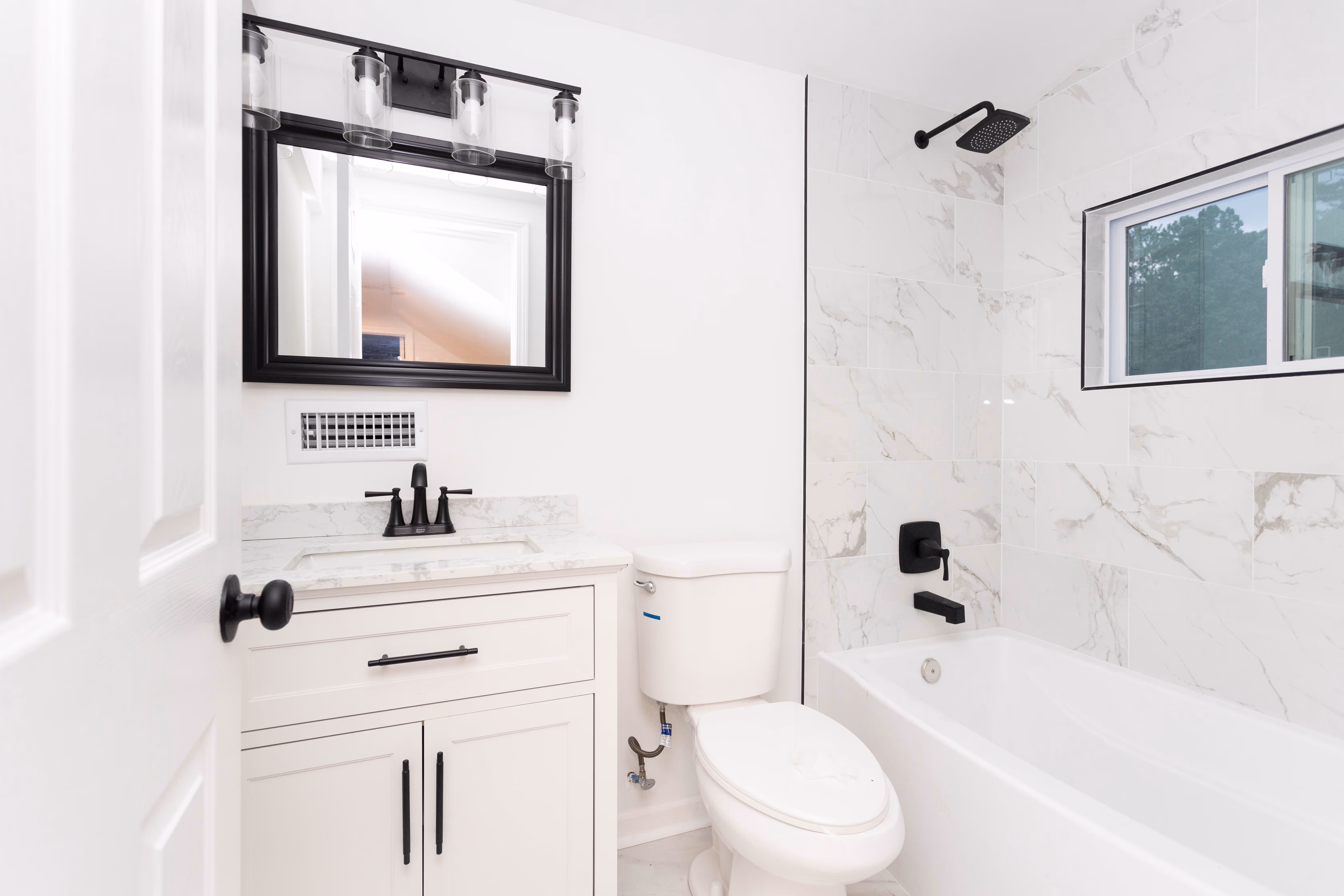 Modern white bathroom with black fixtures, a vanity with quartz countertop, toilet, bathtub, and a window above the tub.