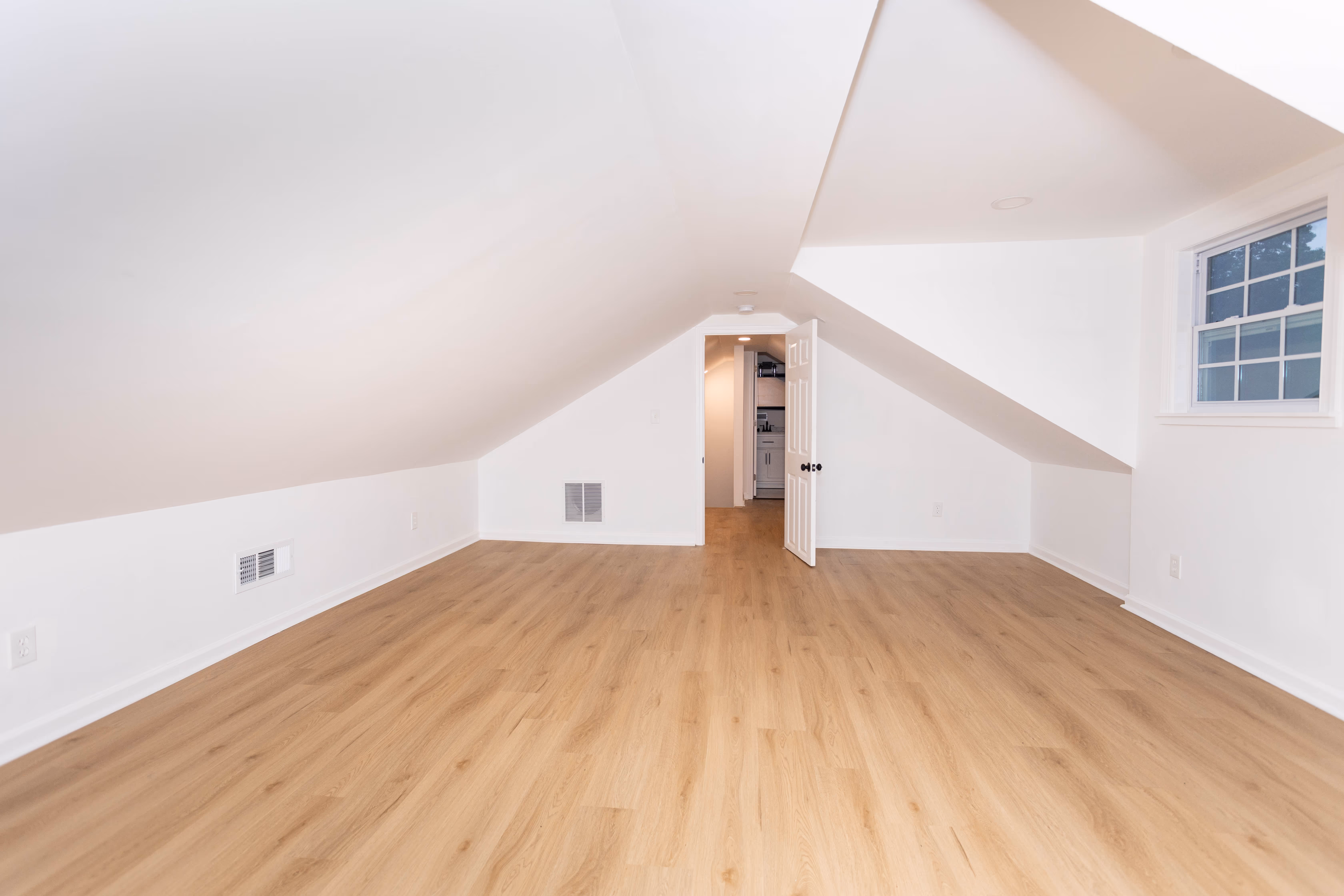 Empty attic room with sloped white walls, light wood flooring, a window, and an open door leading to a small kitchen area.