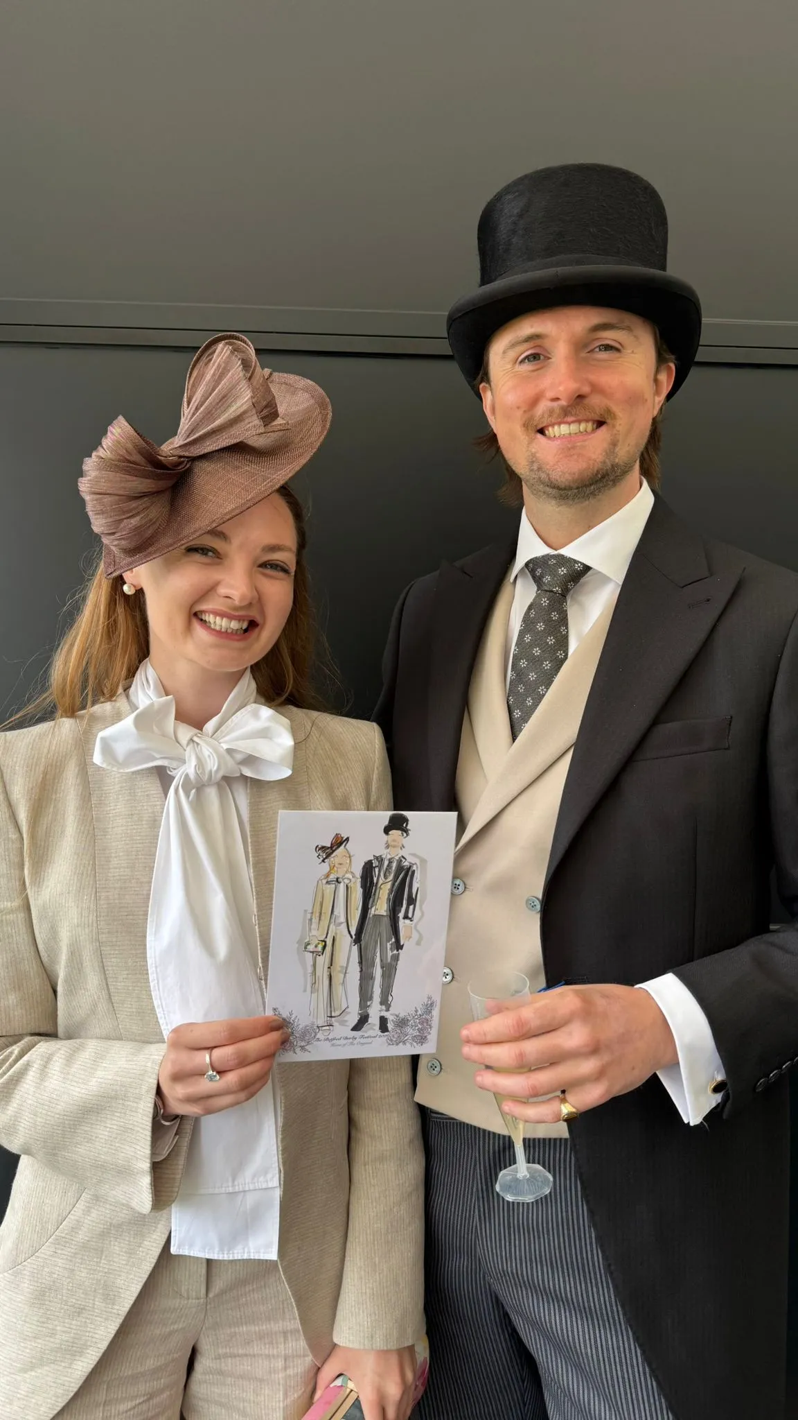 Smiling man and woman in formal attire holding a fashion illustration and a glass of champagne.