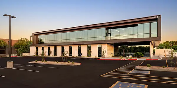 Modern two-story office building with large glass windows, surrounded by a mostly empty parking lot at sunset.
