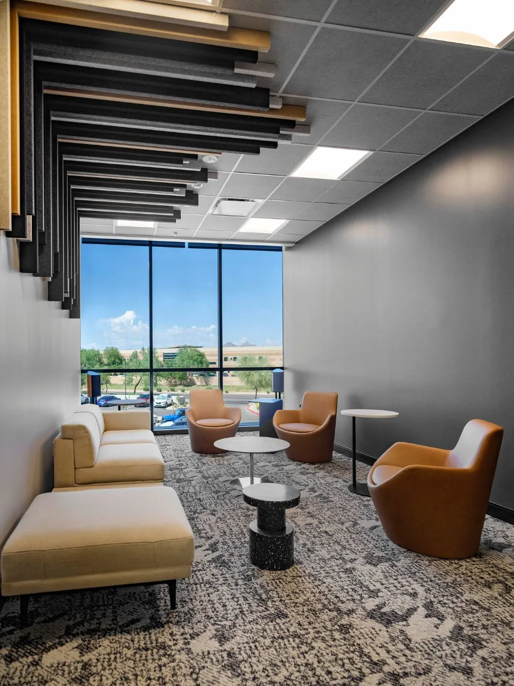 Modern seating area with beige sofa, three brown armchairs, small round tables, and a large window with a view outside.