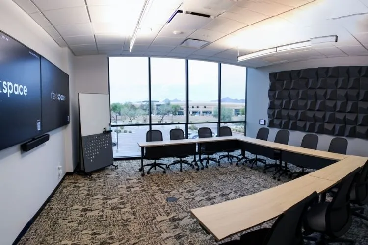 Modern conference room with a U-shaped light wood table, black office chairs, large windows with a city view, a wall-mounted TV screen, and acoustic panels.