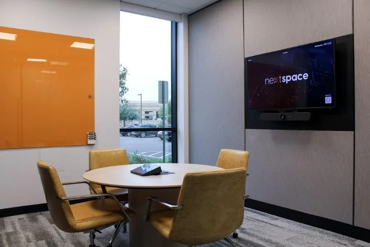 Modern small conference room with round table, four mustard-yellow chairs, orange wall whiteboard, large window, and wall-mounted screen displaying 'nextspace'.
