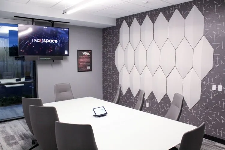 Modern conference room with a white table, six gray chairs, a mounted TV displaying 'nextspace', and geometric wall panels.