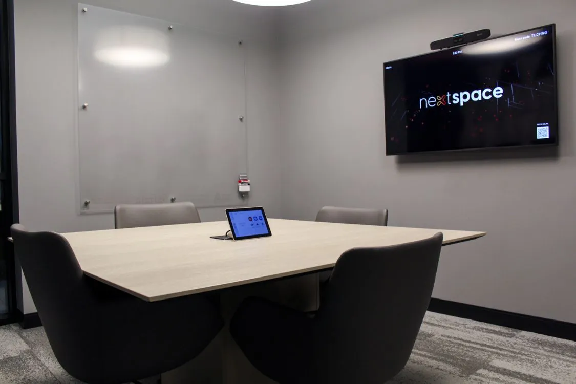 Modern conference room with a rectangular table, four gray chairs, a tablet on the table, a blank whiteboard on the wall, and a TV screen displaying 'nextspace'.