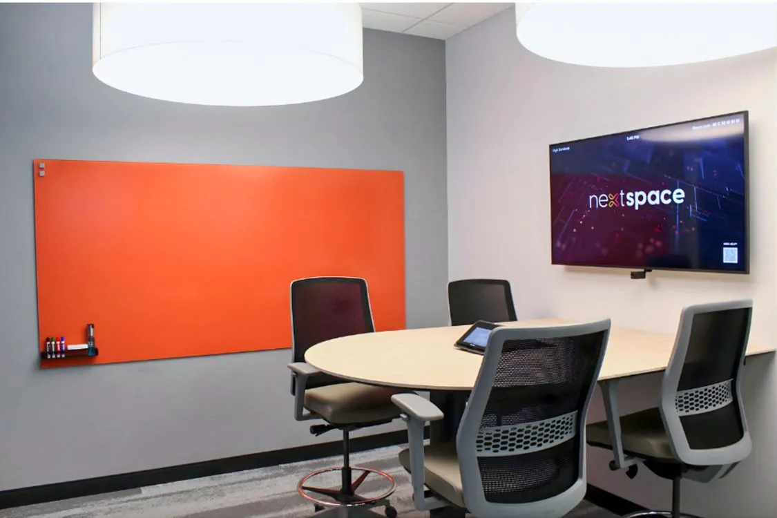 Small modern conference room with round table, four mesh office chairs, an orange whiteboard, and a wall-mounted screen showing the Nextspace logo.