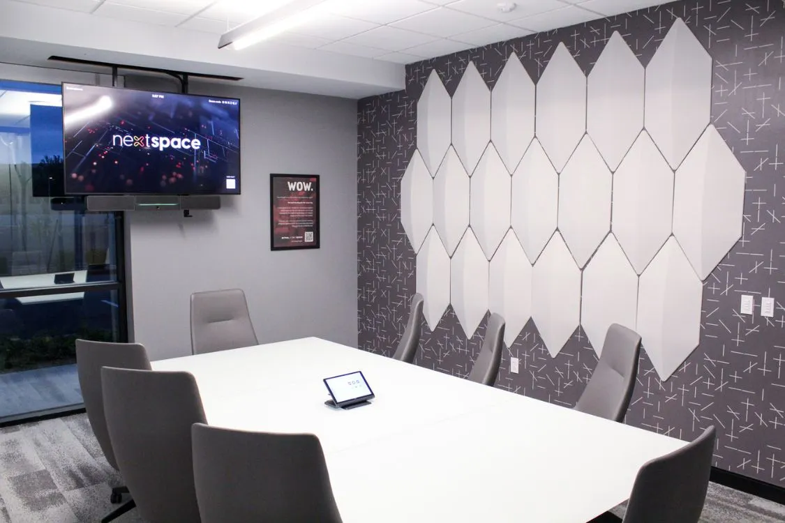 Modern conference room with white rectangular table, gray chairs, geometric wall art, and a TV screen displaying 'nextspace'.