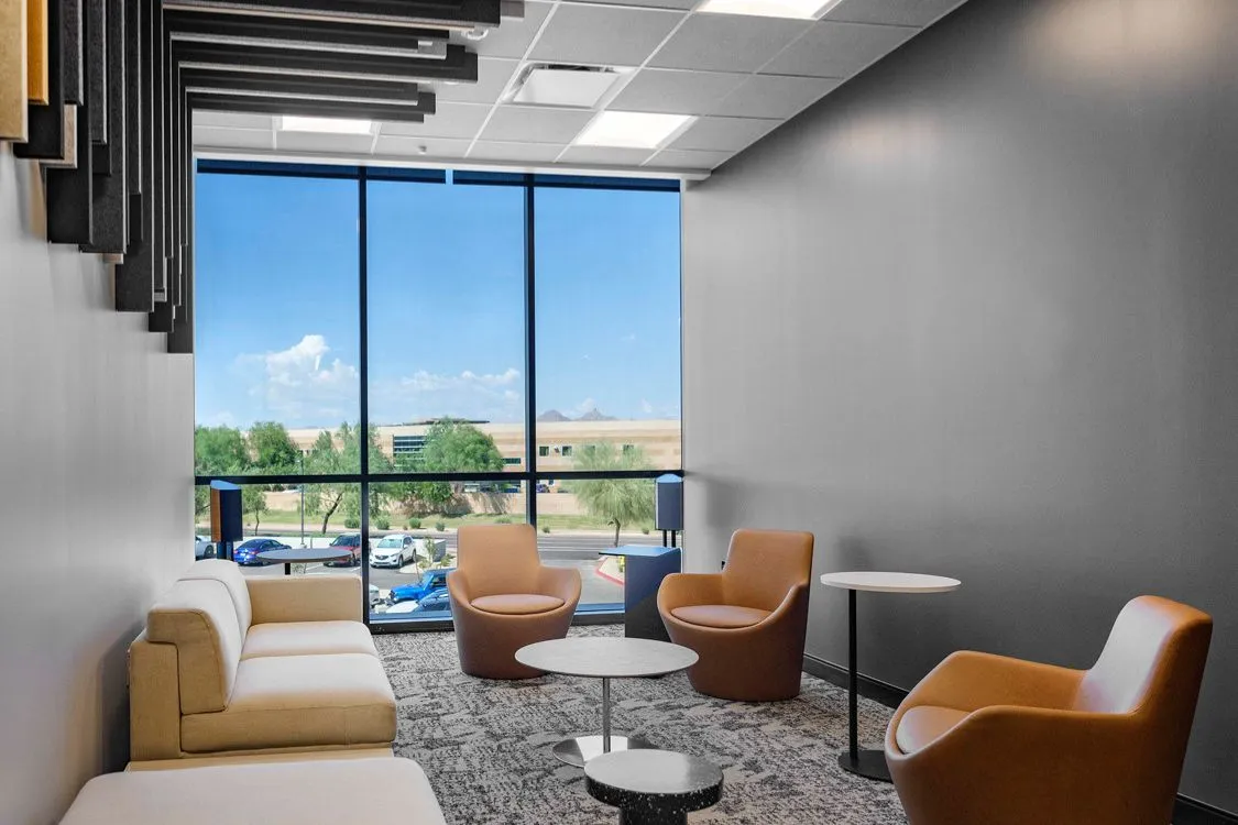 Modern lounge with beige sofa, three brown armchairs, white and black tables, patterned carpet, and large window overlooking parking lot.