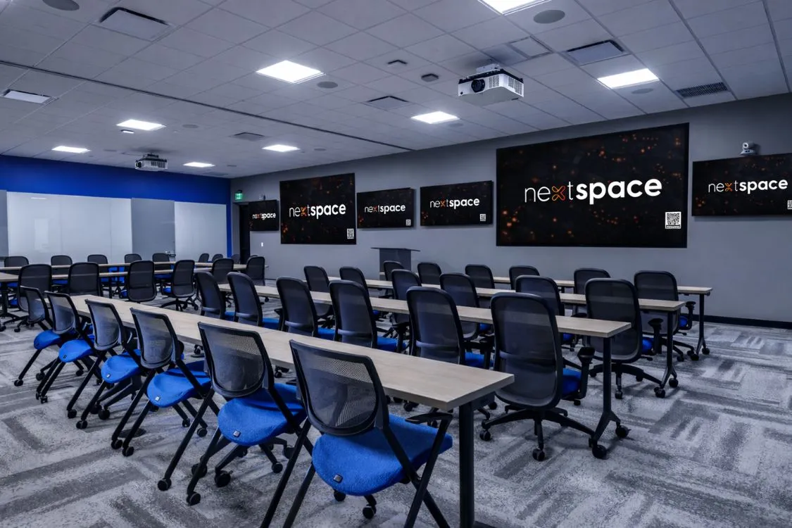 Modern training center with rows of tables and black mesh chairs with blue seats, multiple screens on the wall displaying 'nextspace' logo.