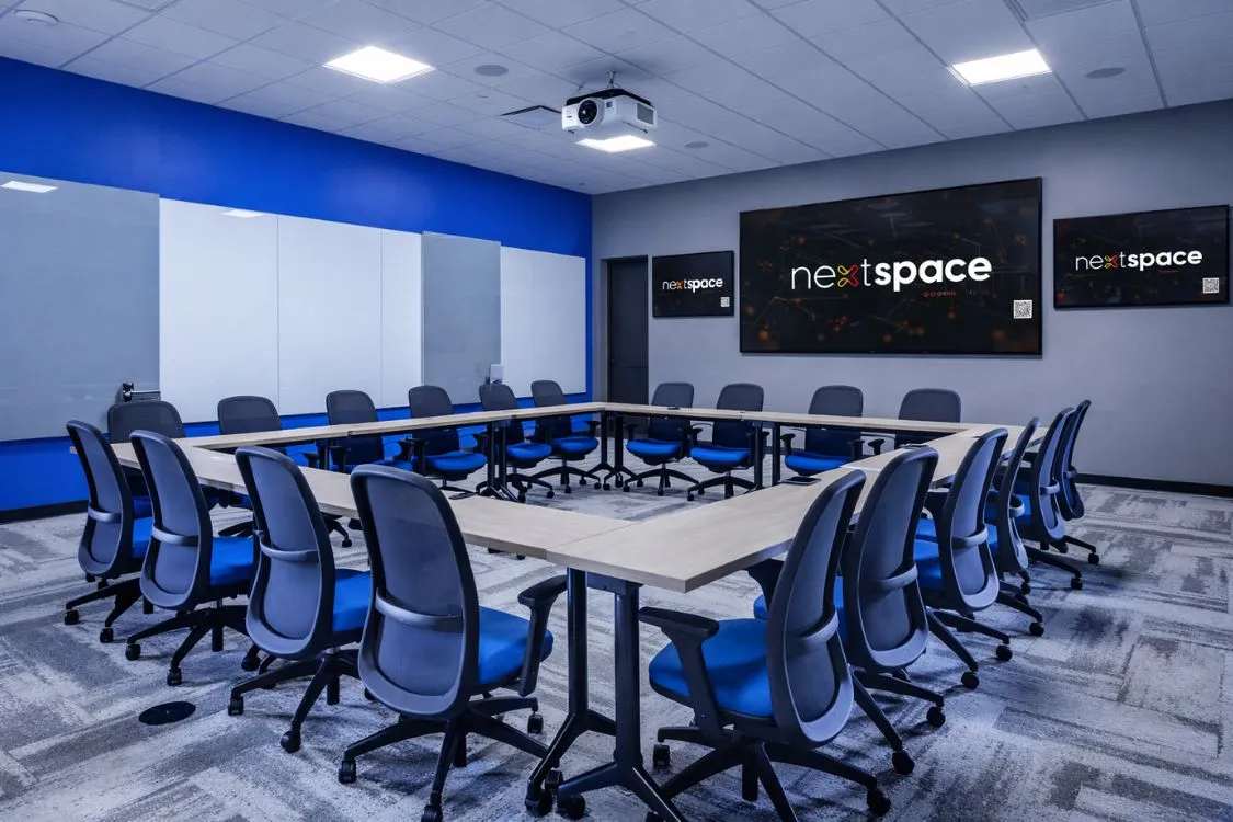 Modern conference room with U-shaped wooden tables and blue ergonomic office chairs, featuring three screens displaying the 'nextspace' logo on the wall.