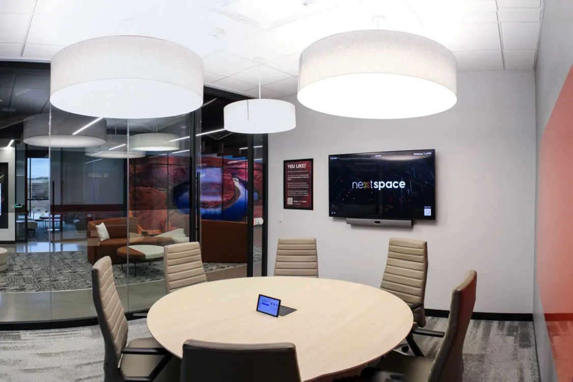 Modern conference room with a round table, six beige chairs, large overhead lights, and a wall-mounted screen displaying 'nextspace'.