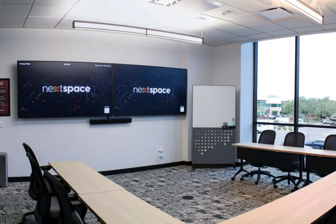 Modern conference room with a long white table, gray upholstered swivel chairs, large hanging circular lights, and two large screens displaying 'nextspace'.