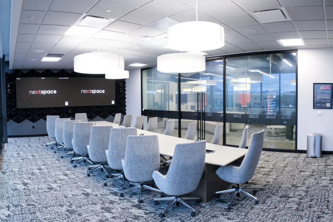Modern conference room with a long white table, gray upholstered swivel chairs, large hanging circular lights, and two large screens displaying 'nextspace'.