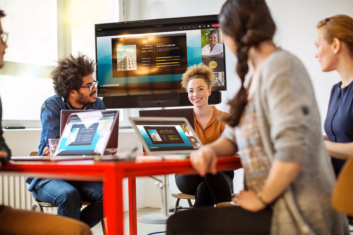 Five coworkers in a modern office having a virtual meeting with laptops and a screen displaying a presentation about historic renovation of an old city library.