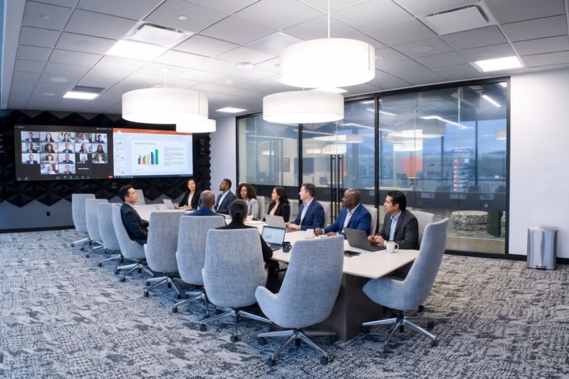 Modern conference room with a long white table surrounded by gray swivel chairs, large ceiling lights, and dual screens displaying 'nextspace'.